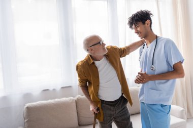 Smiling senior man with walking cane looking at african american nurse at home 