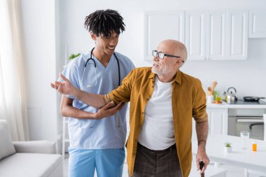 Positive african american nurse in uniform looking at pensioner with walking cane at home 