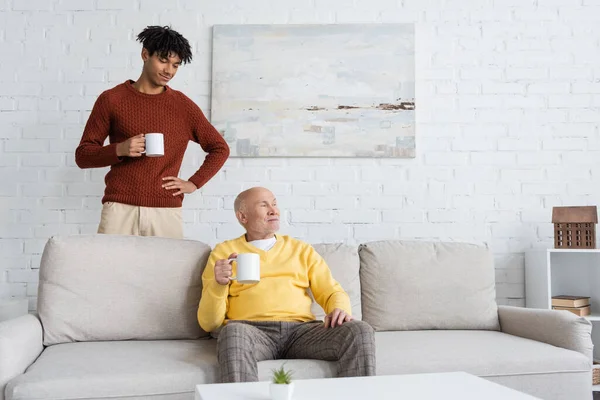 African American Grandson Holding Cup Grandpa Sitting Couch Home — Stockfoto