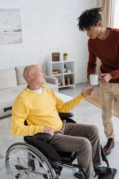 African American Grandson Holding Cup Grandpa Wheelchair Home — Stock Photo, Image