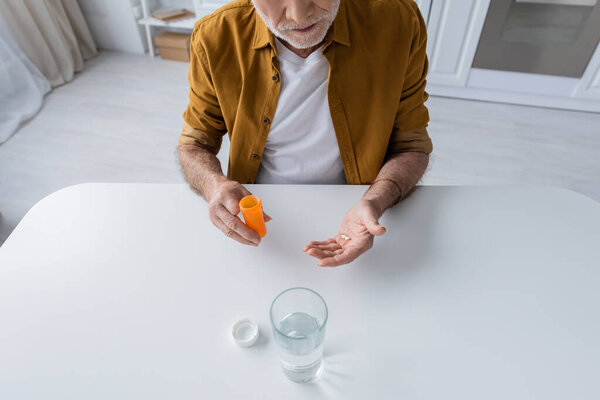 Cropped view of elderly man holding pills near glass of water in kitchen 