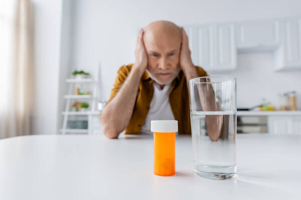 Pills and glass of water near blurred pensioner at home 