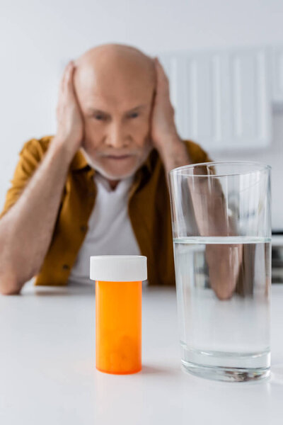 Glass of water and pills near blurred elderly man at home 