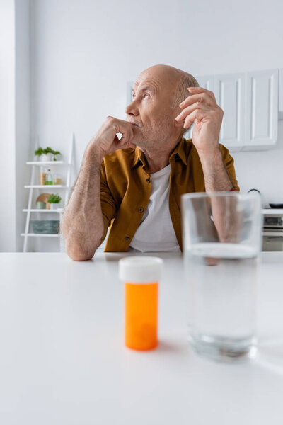 Elderly man looking away near blurred pills and water on table at home 