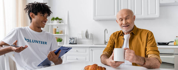 Cheerful african american volunteer holding clipboard near elderly man with cup in kitchen, banner 
