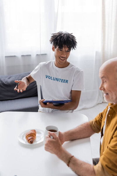 Cheerful african american man holding clipboard near blurred pensioner with cup at home 
