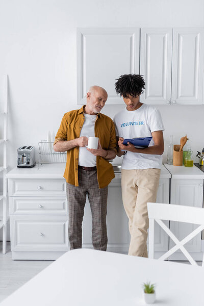 Elderly man holding cup near african american volunteer with clipboard in kitchen 
