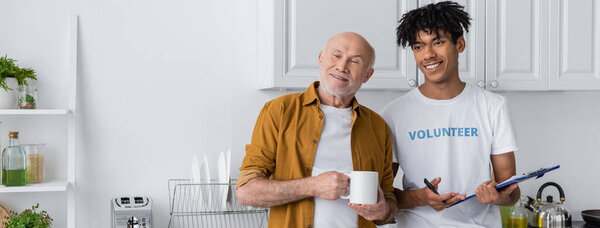 Cheerful african american volunteer holding clipboard near pensioner with cup in kitchen, banner 