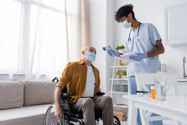Senior man in medical mask sitting in wheelchair near african american nurse with pyrometer and digital tablet in kitchen