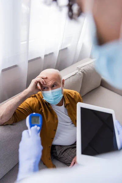 Senior man in medical mask sitting near blurred african american nurse with digital tablet and pyrometer at home