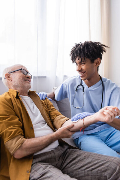 Улыбающаяся африканская американская медсестра держит вату на руках у пожилого пациента дома 