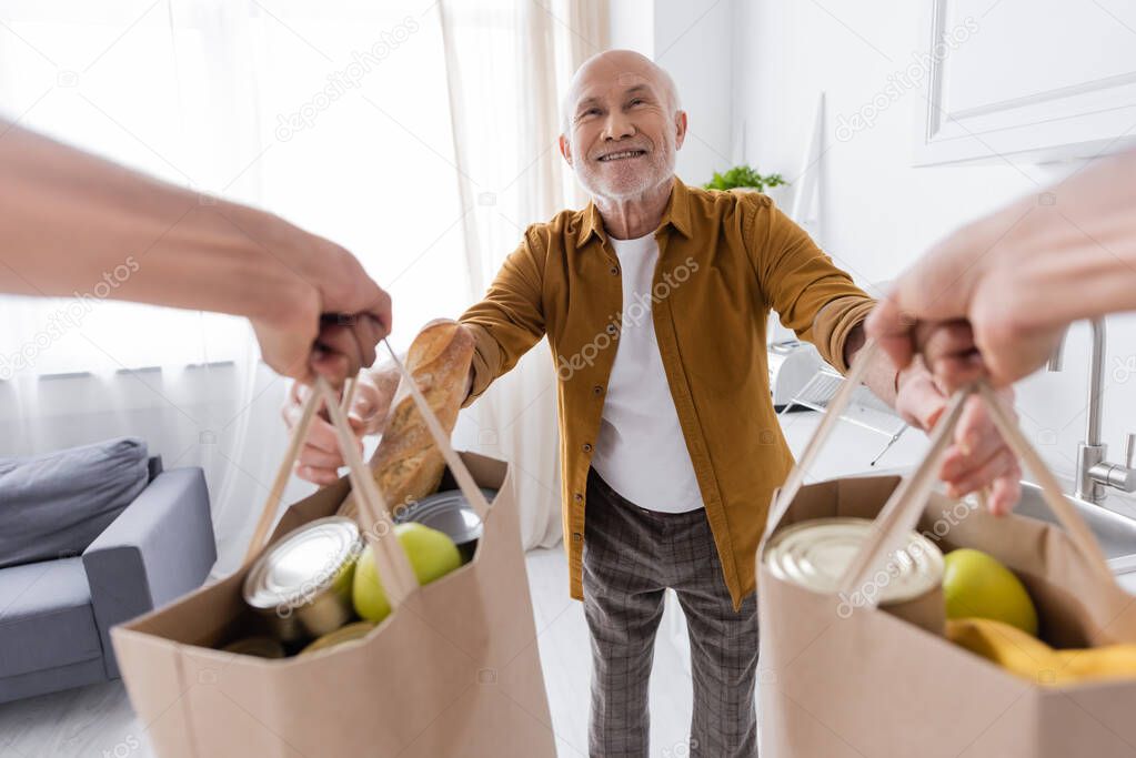 Cheerful pensioner taking bags with food from african american volunteer at home 