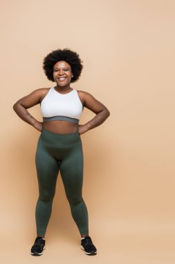 full length of happy african american plus size woman in crop top and leggings posing with hands on hips on beige
