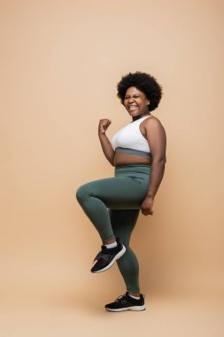 full length of happy african american plus size woman in sportswear showing yes gesture on beige