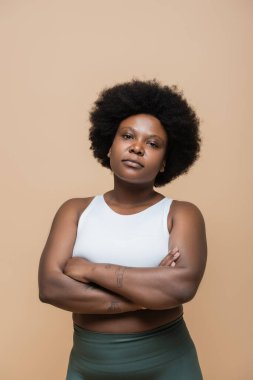 curly african american plus size woman in crop top standing with crossed arms isolated on beige