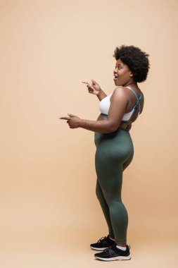 full length of amazed african american plus size woman in sportswear pointing with fingers on beige