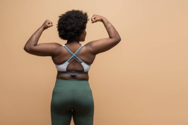 back view of tattooed african american plus size woman in crop top showing power gesture isolated on beige