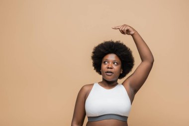 curly african american plus size woman with raised hand pointing with finger isolated on beige
