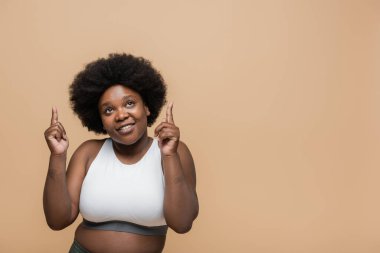 cheerful african american plus size woman in crop top pointing with fingers isolated on beige