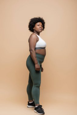 full length of curly african american plus size woman in crop top and leggings standing on beige