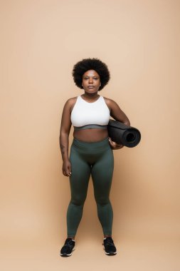 full length of african american plus size woman in crop top holding fitness mat on beige