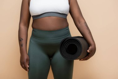 cropped view of tattooed african american plus size woman holding fitness mat isolated on beige 