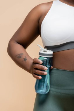cropped view of tattooed african american plus size woman holding sports bottle isolated on beige