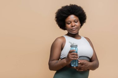 curly african american plus size woman in crop top holding sports bottle with water isolated on beige