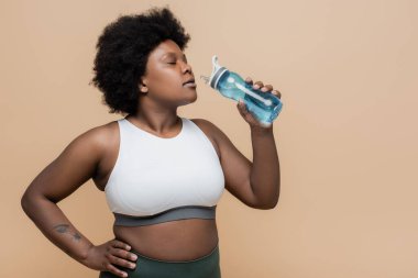 african american plus size woman holding sports bottle with water and standing with hand on hip isolated on beige