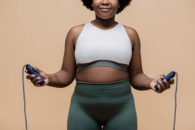 cropped view of cheerful african american plus size woman in sportswear holding skipping rope isolated on beige 