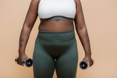 cropped view of tattooed african american plus size woman in sportswear holding dumbbells isolated on beige 