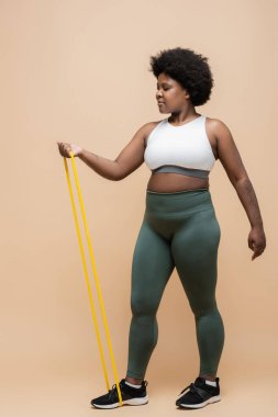 full length of curly african american plus size woman in sportwear exercising with resistance band on beige