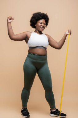 full length of joyful african american plus size woman in sportwear exercising with resistance band on beige