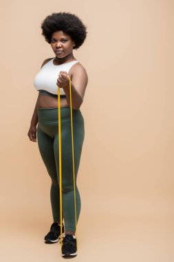full length of african american plus size woman in sportwear exercising with resistance band on beige