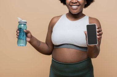 cropped view of happy african american plus size woman holding sports bottle and smartphone with blank screen isolated on beige