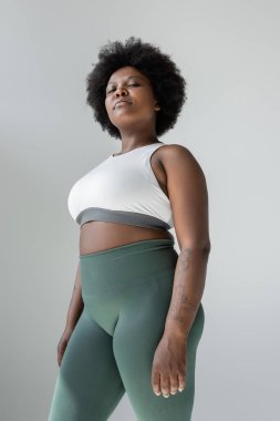 low angle view of tattooed african american plus size woman in sportswear standing isolated on grey