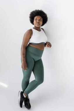 full length of curly african american plus size woman in sportswear standing near white wall 
