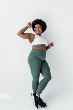 full length of cheerful african american plus size woman in sportswear standing near white wall 