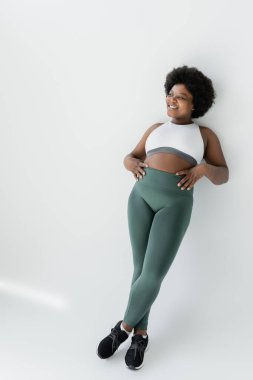 full length of positive african american plus size woman in sportswear standing near white wall 