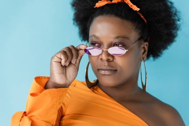 curly african american plus size woman in hoop earrings adjusting sunglasses isolated on blue
