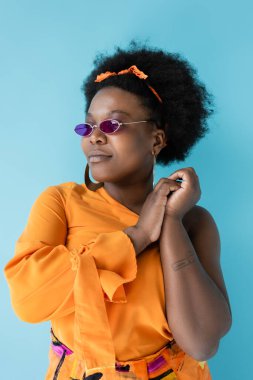curly african american plus size woman in pink sunglasses and hoop earrings isolated on blue