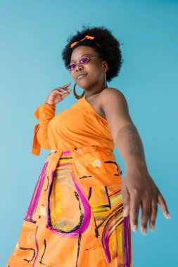curly african american plus size woman in sunglasses and hoop earrings gesturing isolated on blue