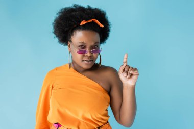 curly african american plus size woman in sunglasses and hoop earrings pointing up isolated on blue
