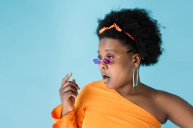 shocked african american body positive woman in pink sunglasses isolated on blue