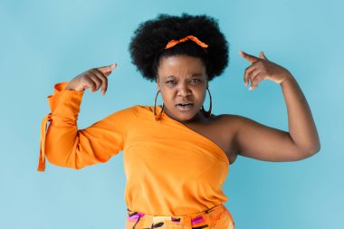 african american body positive woman in orange dress pointing with fingers at curly hair isolated on blue
