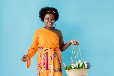 happy african american body positive woman holding straw bag with flowers isolated on blue