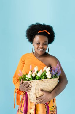 smiling african american body positive woman holding straw bag with flowers isolated on blue
