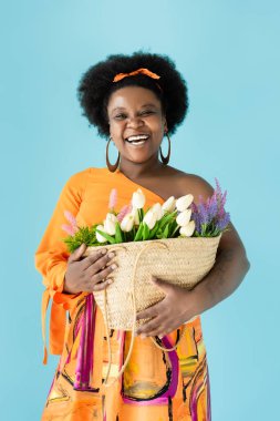 joyful african american body positive woman holding straw bag with flowers isolated on blue