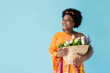 positive african american woman holding straw bag with flowers isolated on blue