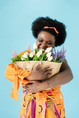 happy african american body positive woman hugging straw bag with flowers isolated on blue